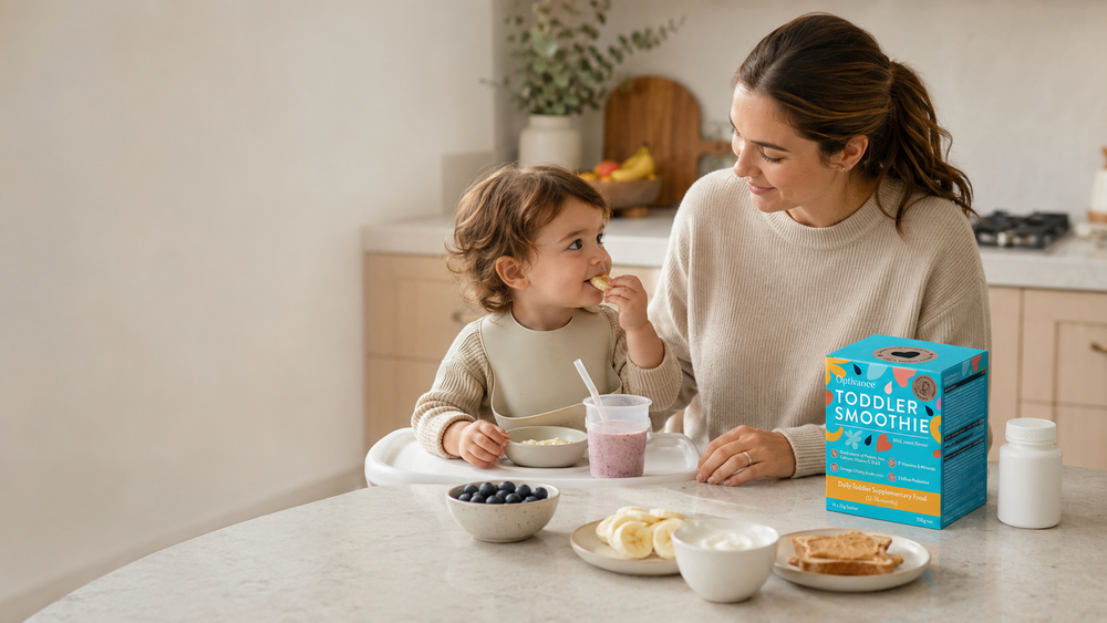 Mum sitting beside her toddler during a calm snack time with Optivance Toddler Smoothie, fruit, yoghurt, toast and a multivitamin bottle on the bench.