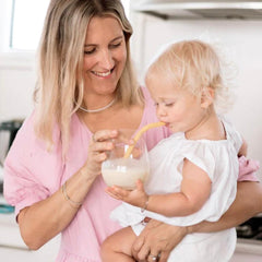 Optivance nutritionist Kristy holding toddler daughter in a bright white kitchen while toddler drinks Optivance Toddler Smoothie through a straw